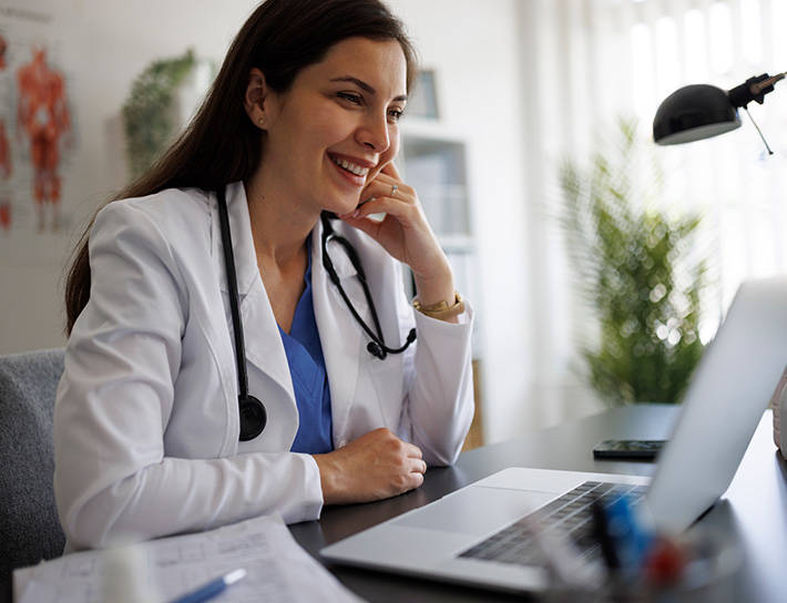 A smiling clinician in a white coat on a video call at her desk